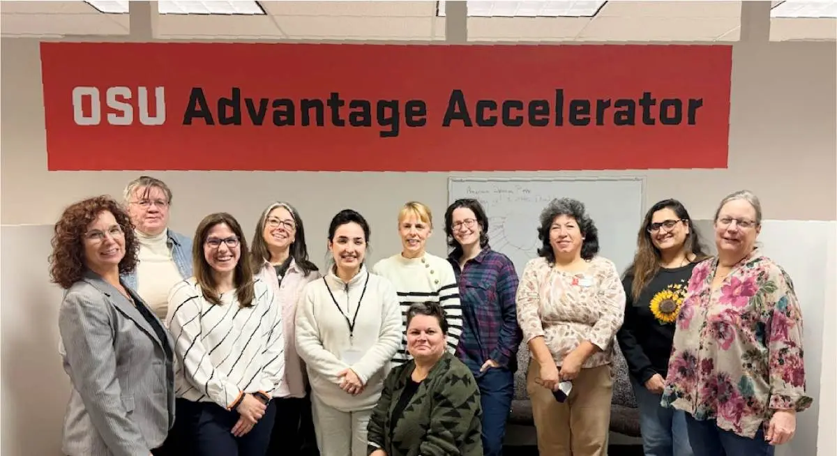 Group photo of eleven people standing and sitting in front of a orange and black 'OSU Advantage Accelerator' sign, smiling at the camera in an indoor office setting.